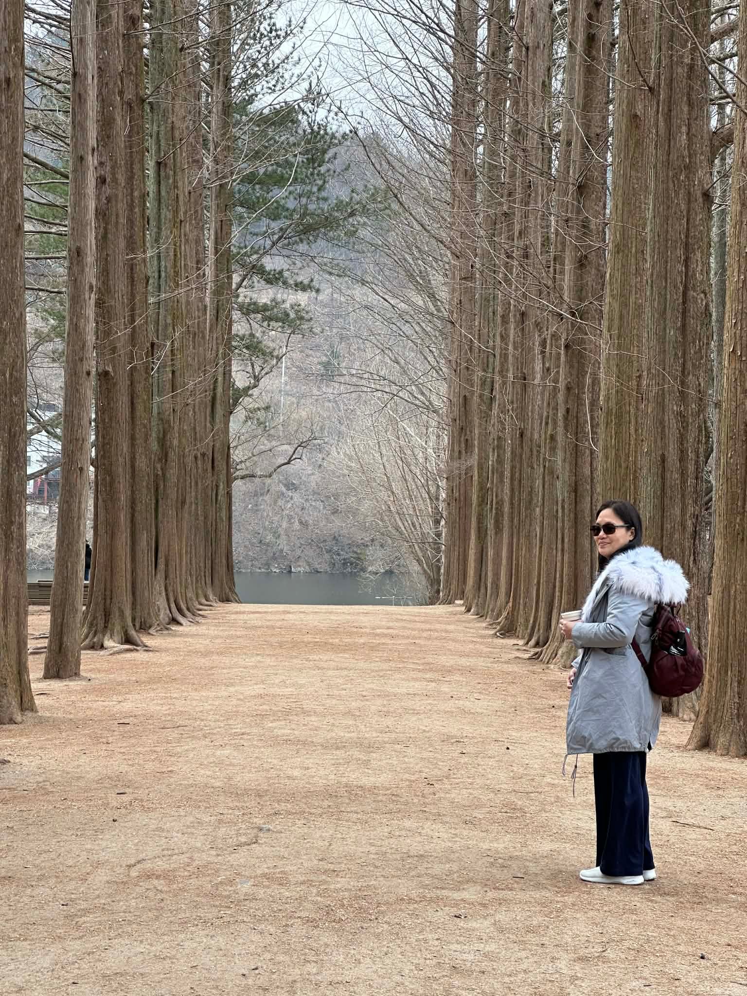 Anissa standing flush to the right of two rows of gigantic trees, a feature that Nami Island is known for. She is wearing a light blue winter coat and shades, looking wistfully behind her. (c) Andro Fernandez